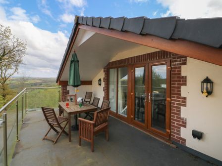 A balcony with wooden table and chairs and a closed green umbrella with glass railing at Scenic Views in High Bickington