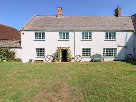 The back exterior of a white two-story house with blue window frames a wooden bench a grassy lawn and a small pergola at Aluric's Hall in Porlock