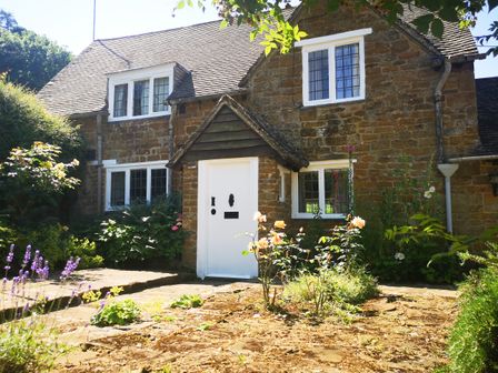 A stone cottage with white framed windows and a white door surrounded by flowers and plants at Spring Cottage in Alkerton near Banbury
