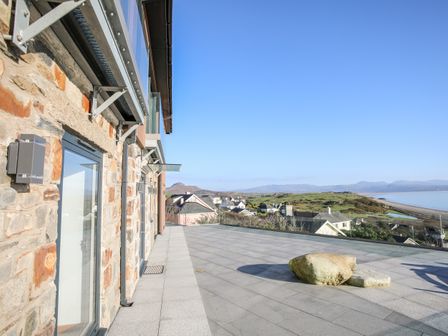 A stone building with glass doors and a tiled terrace overlooking houses hills and a beach at Hafod Y Mor in Criccieth