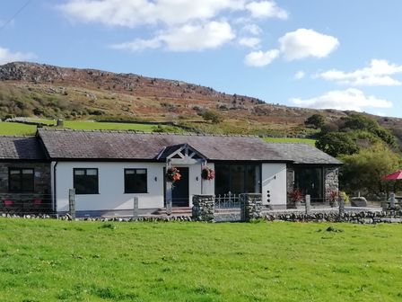 A single story house with a tiled roof and stone and white walls set in a grassy field with hills in the background at Cartref in Trawsfynydd