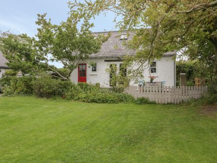 A small white house with a red door surrounded by trees and a lawn at Little House in Red Wharf Bay