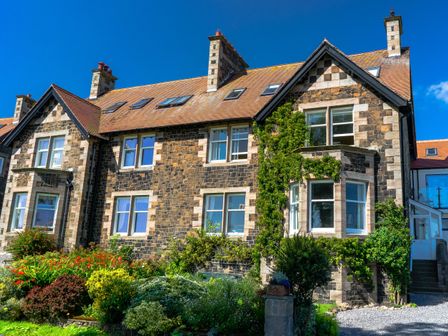 A stone house with multiple windows and chimneys surrounded by bushes and flowers at Whinstones at 4 The Villas in Embleton