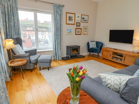 A living room with flowers on a table next to a sofa and armchair near a window and a television on a wooden stand at Grays' Cottage in Whitby
