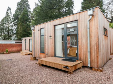 Wooden lodges with glass doors and small wooden steps outside surrounded by gravel and trees at Lakeside Village Lodges & The Potting Shed in Capesthorne near Macclesfield