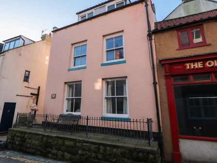A street view of a pink three story house with blue window frames and a bench outside next to a red storefront at Salmon Cottage in Staithes