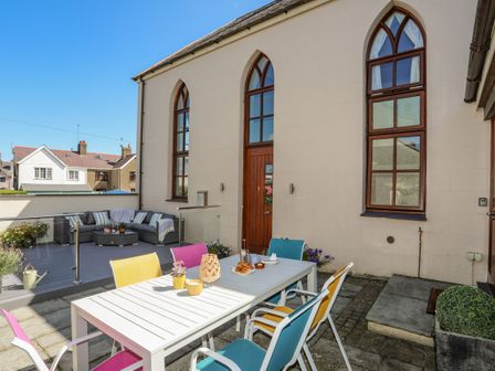 An outdoor patio with a white table and colorful chairs next to a building with arched windows at Capel Tabernacl in Morfa Nefyn