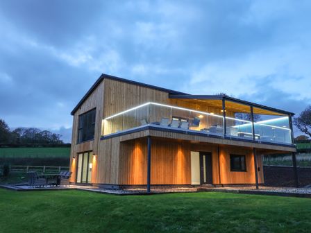 A modern two-story wooden house with an upper balcony and outdoor seating on a lawn at Fron Haul - Robin Goch in Llanfwrog near Ruthin