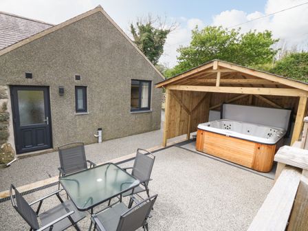 A patio area with a glass table and four chairs next to a wooden shelter housing a hot tub at Tyddyn Sianel - Onnen in Llanystumdwy near Criccieth