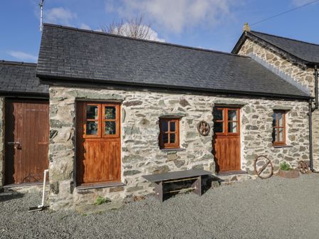 A stone building with wooden doors and windows a bench and metal wheels on the ground at Nyth Y Wennol in Llanfachraeth