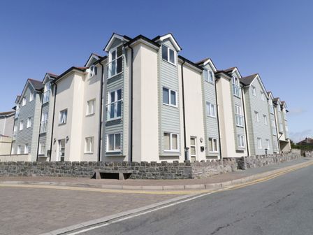 An exterior view of a multi-story residential building with cream and light gray siding along a street at 10 Pen Llanw Tides Reach in Rhosneigr