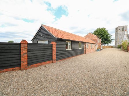 A gravel driveway alongside a black wooden and brick building with a tiled roof near an old stone tower at Cobblers Barn in Repps With Bastwick near Potter Heigham