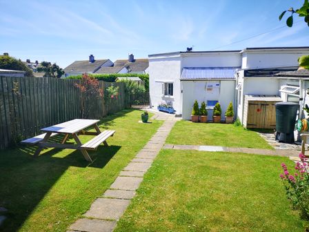 A garden with a wooden picnic table a stone path and potted plants next to a white house with a shed and fence at Gull Cottage in Tintagel