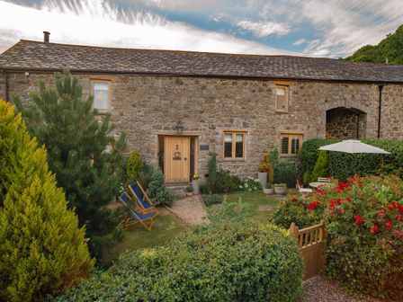 A stone house with wooden door and windows surrounded by garden chairs bushes flowers and a table with umbrella at May Blossom Barn in Lustleigh