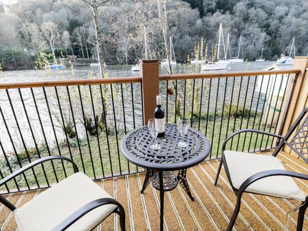 A balcony with a small round metal table with a bottle of wine and two wine glasses and two cushioned chairs overlooking a lake with boats at Macan in Far Sawrey