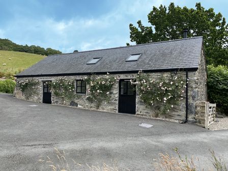 A stone building with two black doors two windows and climbing roses along the boundary at The Shippon in Llanrwst