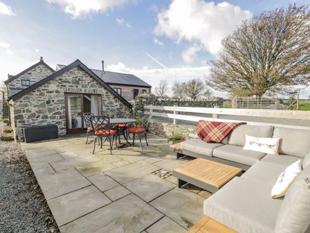 An outdoor patio with a stone building a table with four chairs and a gray sectional sofa at Cwch Gwenyn in Llanfachraeth