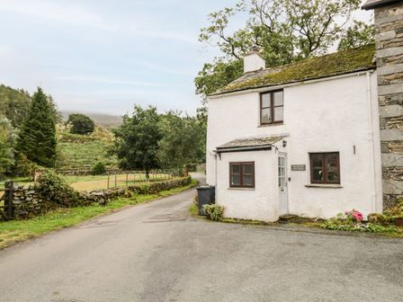 The exterior of a white cottage beside a country road with stone walls and greenery at Beckfold Cottage in Broughton-In-Furness