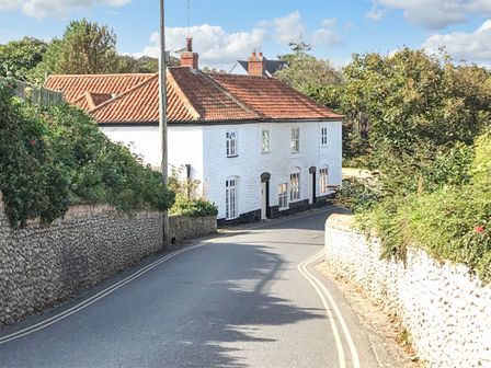 A curved street with white brick walls and a white house with a red tiled roof surrounded by greenery at Cobblers Cottage in Mundesley