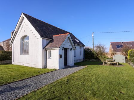 A white building with a slate roof on a grass lawn with a gravel path a picnic table and a shed in the yard at Capel Hermon in Llanynghenedl near Bodedern