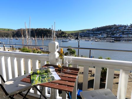 A balcony with a wooden table holding a bottle of wine, two wine glasses and an open magazine overlooking a river with boats and hills in the background at 2 Embankment House Dartmouth