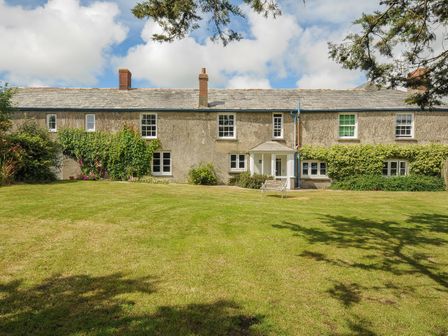 A long stone house with multiple windows and a door with a porch in the center a bench on the lawn in front of the house at Lee Barton Farmhouse in Bude