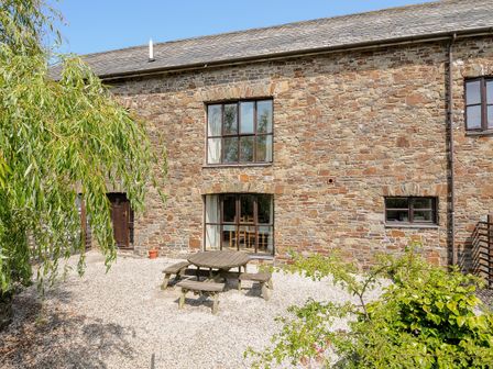An outdoor seating area with a round wooden picnic table and benches outside a stone building with windows at The Shippon in Bude