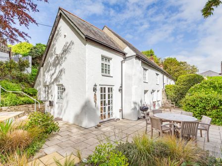A white cottage with a tiled patio having wooden table and chairs and surrounded by bushes at 3 Rose Cottages in St Agnes