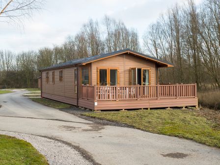 A wooden lodge with a railing around a porch on a grassy plot near a road at Waternook Lodge in South Lakeland Leisure Village
