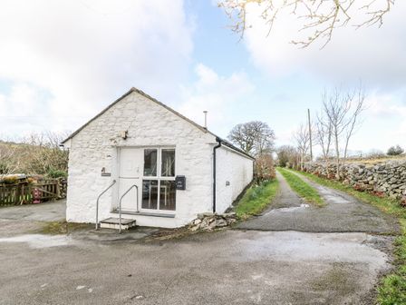 A white stone building with a door and window next to a narrow road with grass and stone walls at Y Buarth in Waunfawr