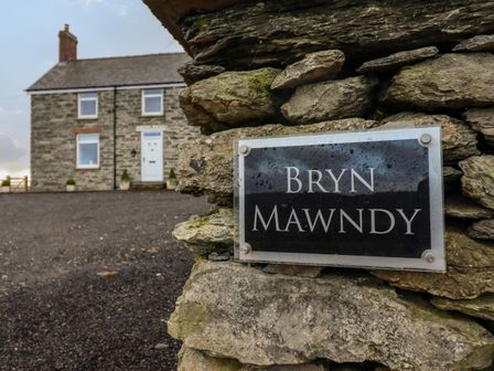 A stone house with a white door and three windows behind a stone wall with a Bryn Mawndy sign at Bryn Mawndy Corwen