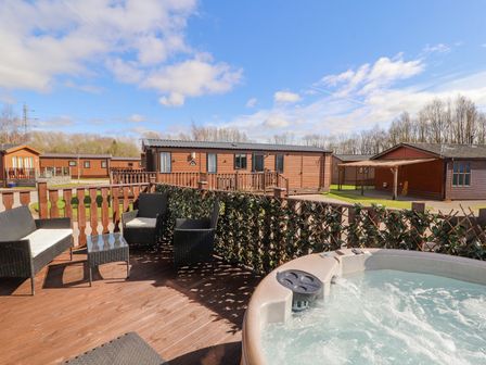 An outdoor deck with a hot tub and wicker chairs surrounded by wooden fences and lodges at Duckling Lodge South Lakeland Leisure Village