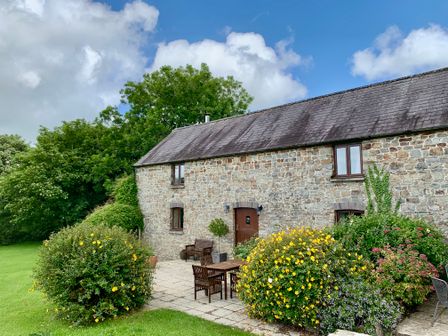A stone building with wooden door and windows surrounded by bushes and outdoor furniture at The Granary in Newport Pembrokeshire