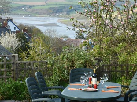 An outdoor dining table set with wine glasses and a bottle of wine on a patio with garden plants and a river in the background at Gellifor in St Dogmaels Pembrokeshire