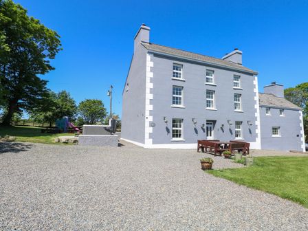 A large gray house with multiple windows and chimneys with outdoor benches and tables on a gravel area at Newton West Farm in Newton near Newgale