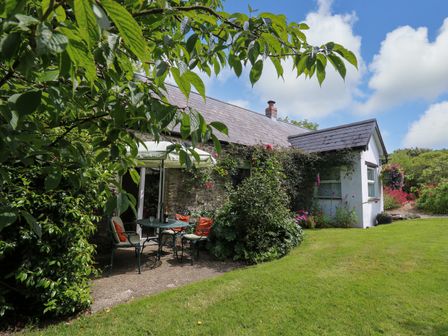 An outdoor seating area with table and chairs under an umbrella beside a stone house with a slate roof and garden plants at Trawsnant Cottage in New Quay Ceredigion