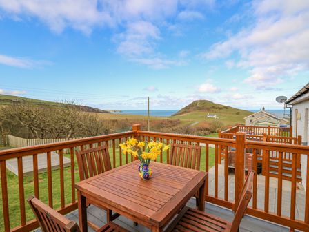 A wooden outdoor table with four chairs and a vase of yellow flowers on a deck overlooking grassy fields and hills by the sea at Llaethdy in Cardigan