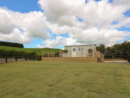 A small building on a wooden deck with flower baskets and fenced grassy area at Marches Way in Rhayader