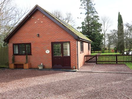 A brick house with a brown door and windows a gravel driveway and a wooden fence with trees and grass in the background at Byefield Lodge Welland
