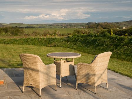 A patio with two wicker chairs and a round table overlooking green fields and hills at Tyddyn Haidd in Amlwch