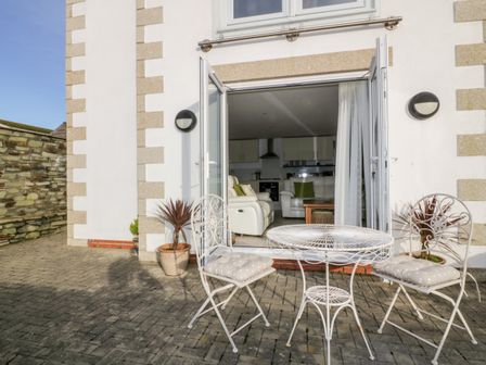 An outdoor patio with a white metal table and two chairs with cushions outside a building with open double doors showing a living room with white sofas at 3 Spyrys Heyl in Newquay