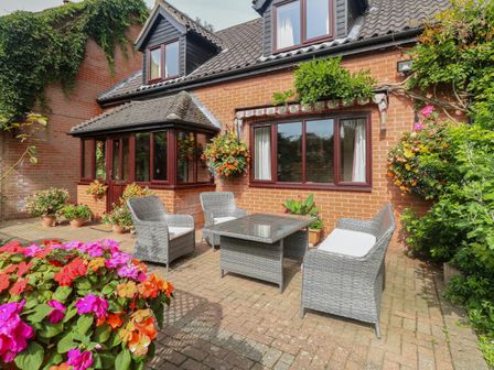 A patio with outdoor wicker furniture and cushions surrounded by flower pots and hanging plants at Walnut Tree Cottage in Catfield