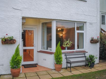 The exterior of a white house with a glass door two potted plants a bench and two hanging flower baskets at Fairview in Salcombe