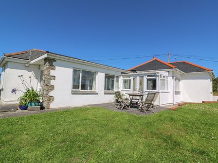 A white bungalow with a patio table and chairs on a paved area and a grassy lawn at Rospletha Bungalow in Treen