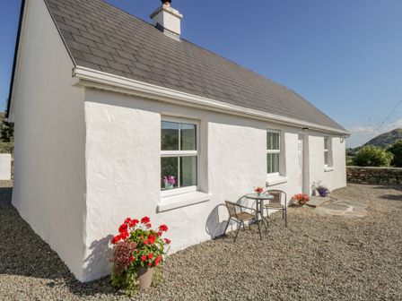 An exterior view of a white cottage with a slate roof and gravel ground with potted flowers and a small table with two chairs outside at Tigh Mhicheal Phaidin Cloghbrack Middle near Clonbur County Galway