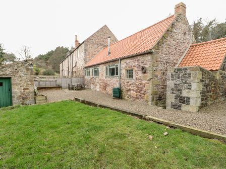A stone house with a tiled roof a green door a gravel patio with outdoor furniture and a grassy lawn at The Old Workshop in Middleton near Belford