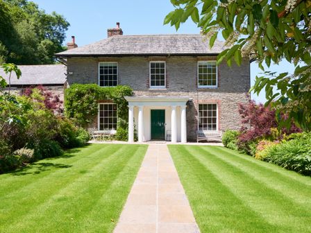 A stone house with white columns green door and manicured lawn with garden benches and plants at Gitcombe House in Cornworthy