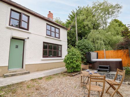 An outdoor patio area with a wooden table and chairs a hot tub and a light green door on a white house at Fisherman's Cottage in Llandysul