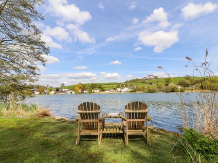 Two wooden chairs and a small table on grass facing a lake with houses and green hills in the background at Clerk Beck Cottage in Great Urswick near Ulverston