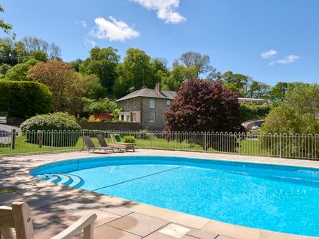 An outdoor swimming pool with lounge chairs and a fence with a house and trees in the background at Garden House in Cornworthy